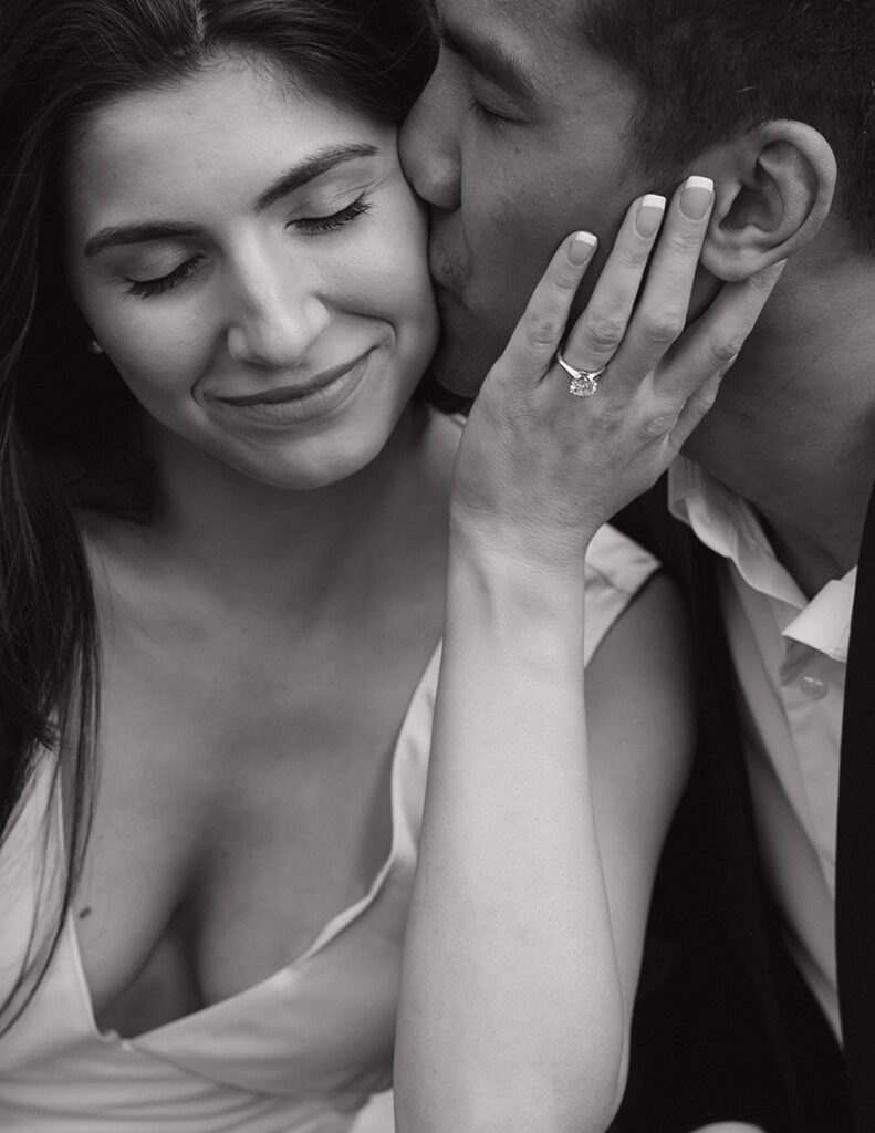 A black and white close-up of a couple sharing an intimate moment. The man gently kisses the woman's cheek. Her hand, displaying a ring, rests on his face. Both exhibit serene, content expressions.