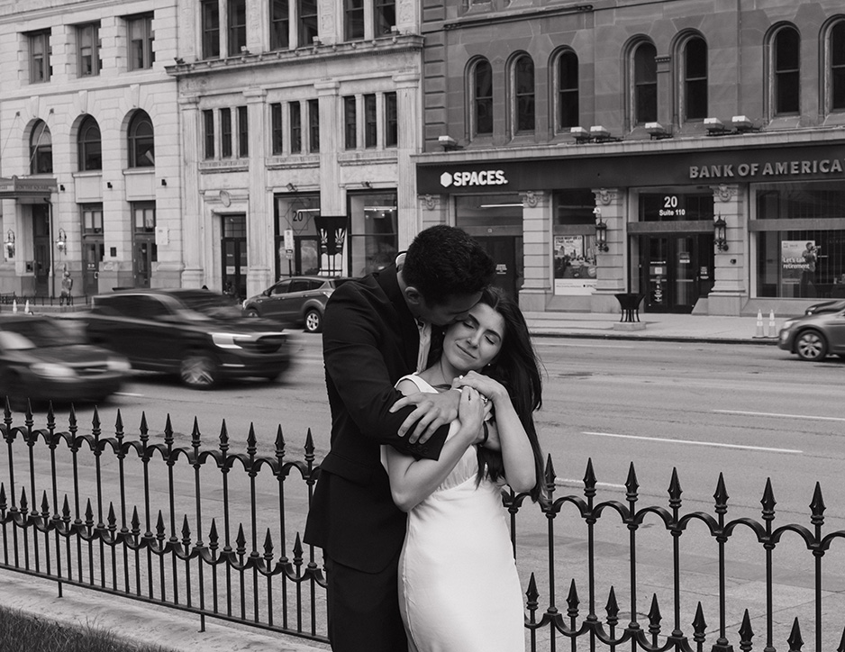 A couple embraces affectionately beside a wrought iron fence on a busy city street, with historic buildings in the background. The scene feels romantic and intimate.