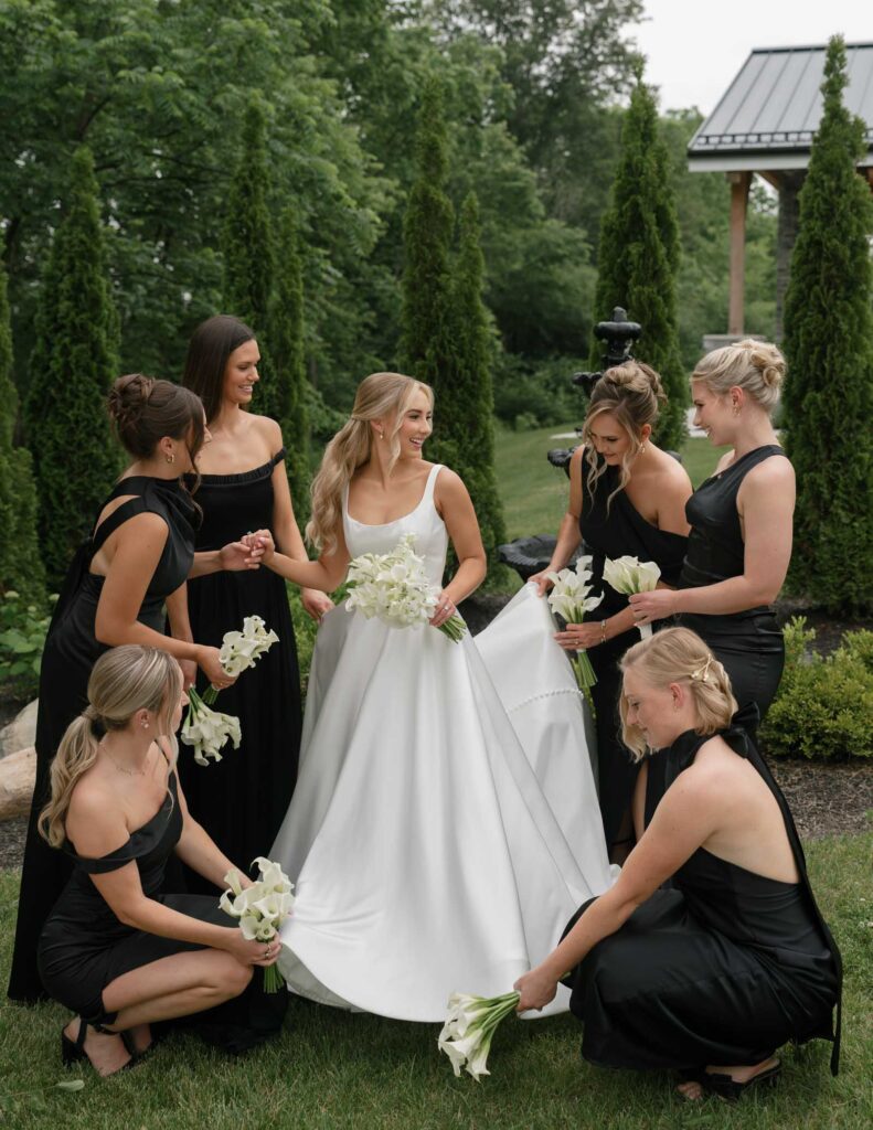 An Ohio bride in a white gown is surrounded by bridesmaids in black dresses holding white flowers, set against lush greenery in a garden.