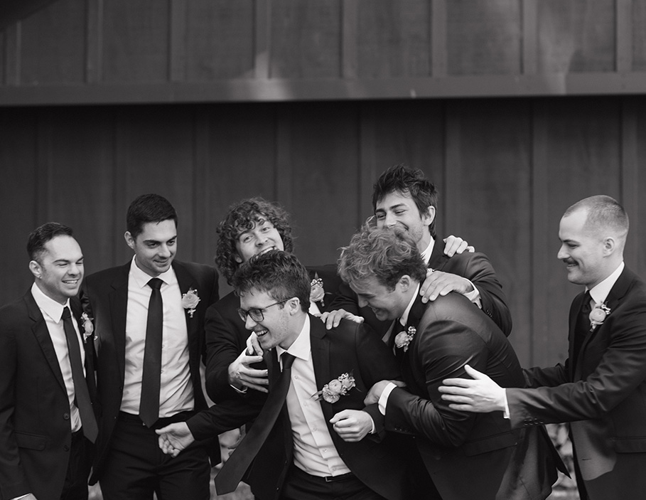 A group of seven groomsmen in suits share a joyful moment, laughing and embracing. The black-and-white setting conveys camaraderie and celebration.