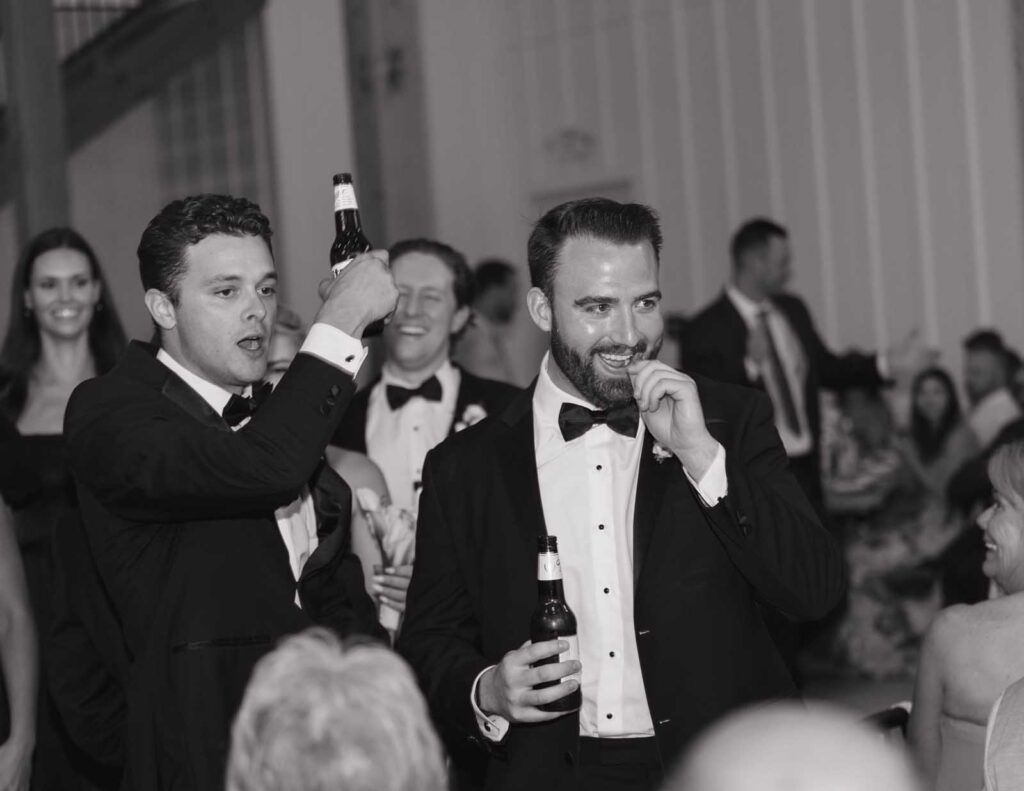 A group of men in tuxedos, smiling and holding beer bottles, appear to be celebrating. The atmosphere is joyful and lively in a formal setting.