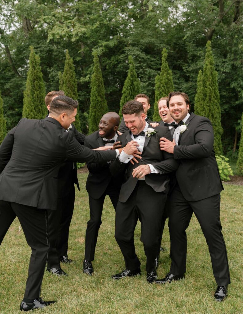 A group of groomsmen in black suits and bow ties joyfully embrace and laugh in a garden setting, surrounded by lush greenery and tall shrubs.
