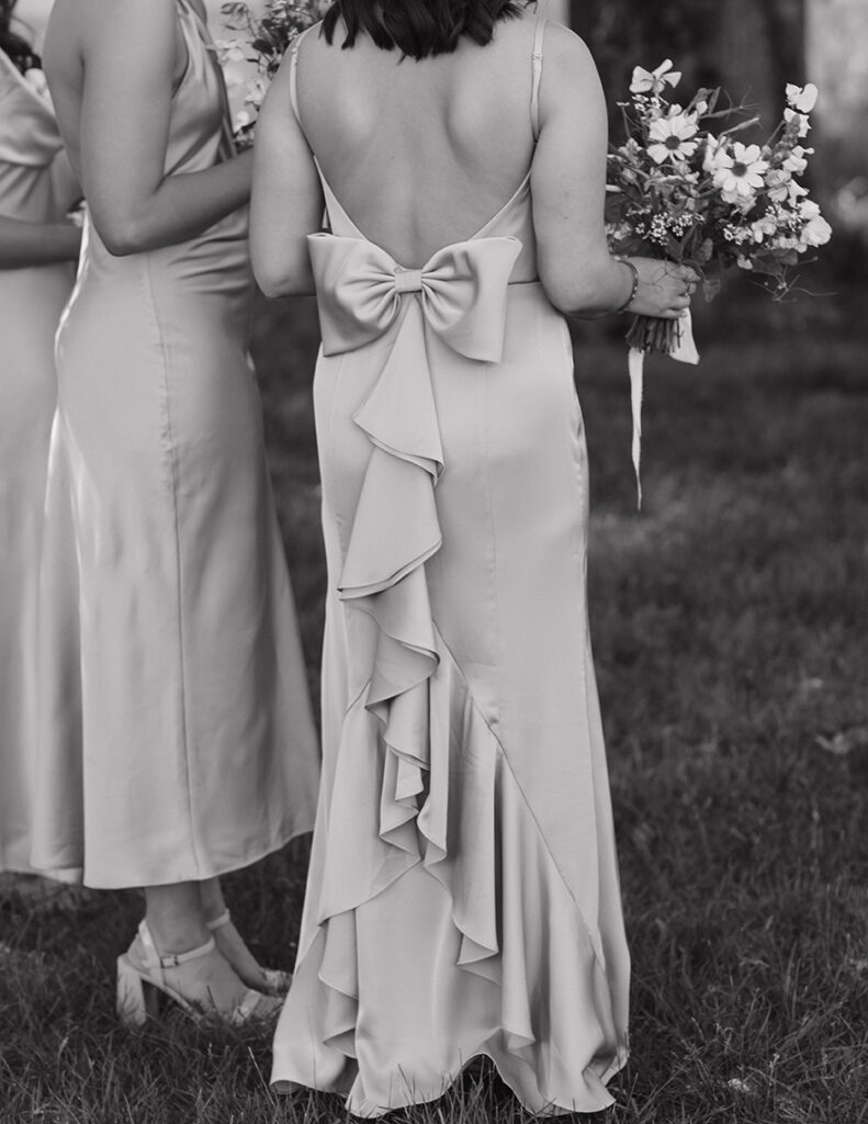 Two women in elegant satin dresses stand on grass, holding bouquets. One has a large bow and ruffles on the back. The scene feels serene and formal.