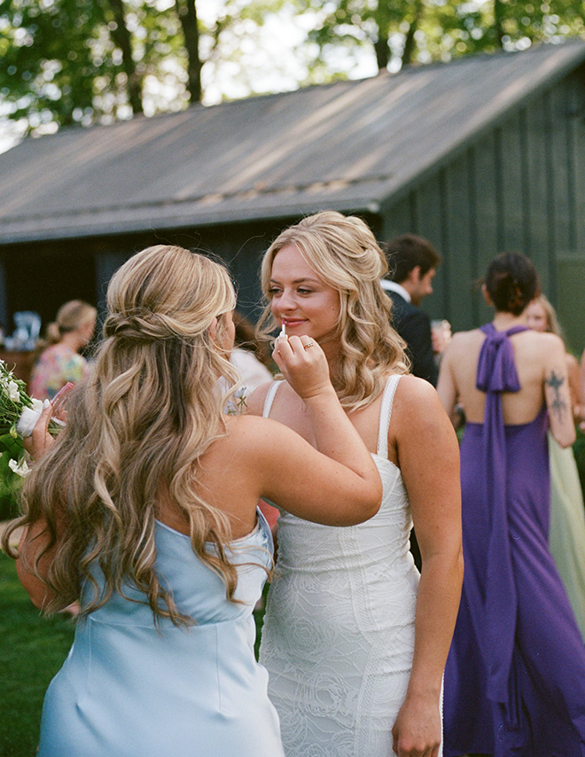 Two women with long, wavy hair stand close together at an outdoor event. One adjusts the other's lipgloss, both smiling softly, with a lively gathering in the background.