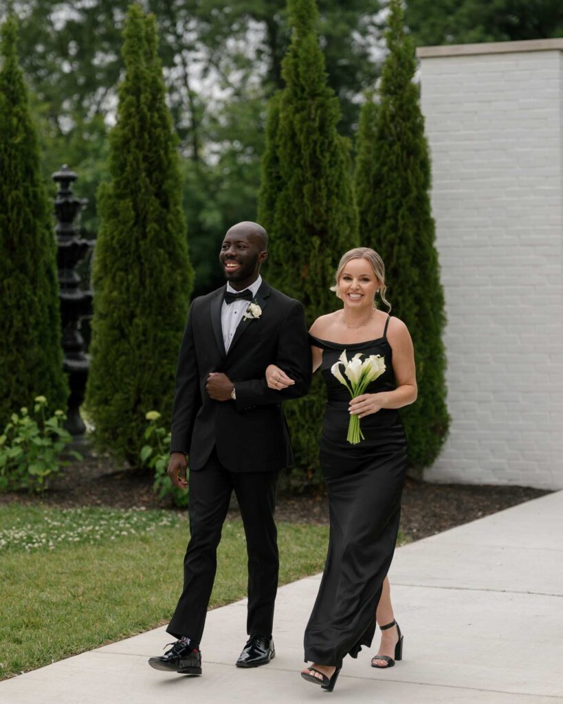 A smiling man in a tuxedo and woman in a black gown walk arm-in-arm on a garden path. The woman holds a bouquet of white flowers.