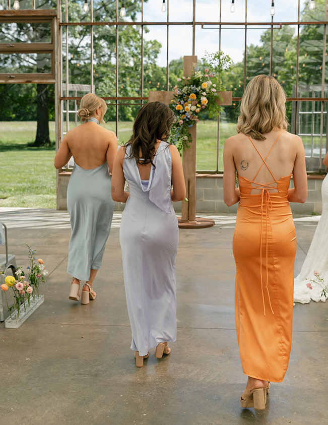 Three women in elegant pastel dresses walk towards a floral altar in a glass-walled venue. The mood is serene and the setting is lush and green.