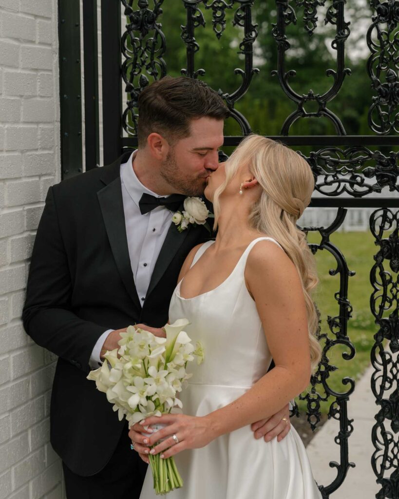A newlywed couple kisses joyfully. The groom is in a black tuxedo, and the bride wears a white dress, holding a bouquet of white flowers. Romantic atmosphere.