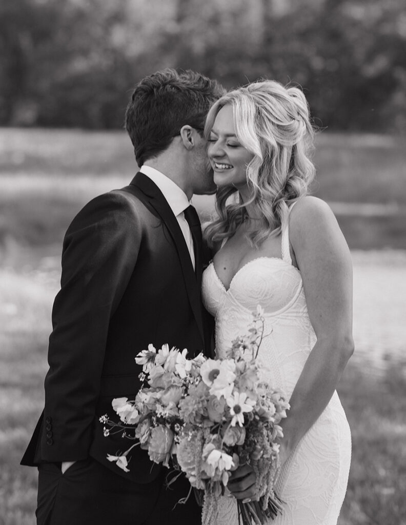Bride in a lace dress smiles joyfully while holding a bouquet as the groom whispers in her ear, set against a serene outdoor backdrop. Black and white.