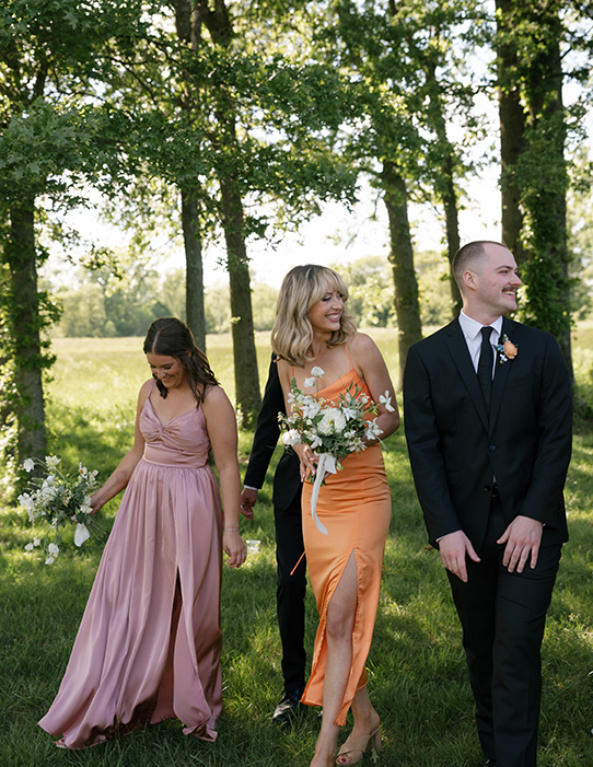 A joyful group of two women in colorful dresses and two men in suits walk through a sunlit forest clearing, holding white floral bouquets.