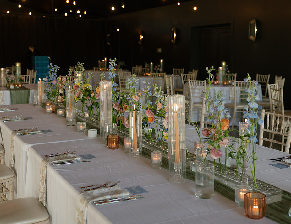 Elegant banquet setting with long tables adorned with delicate blue and pink flowers in glass vases, and flickering candles, creating a cozy ambiance.