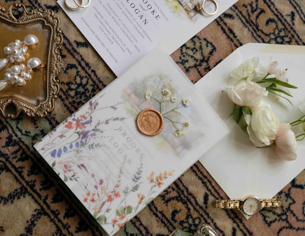 Elegant flat lay featuring a floral-themed wedding invitation with a wax seal, an envelope with white flowers, pearl jewelry, a watch, and wedding rings.