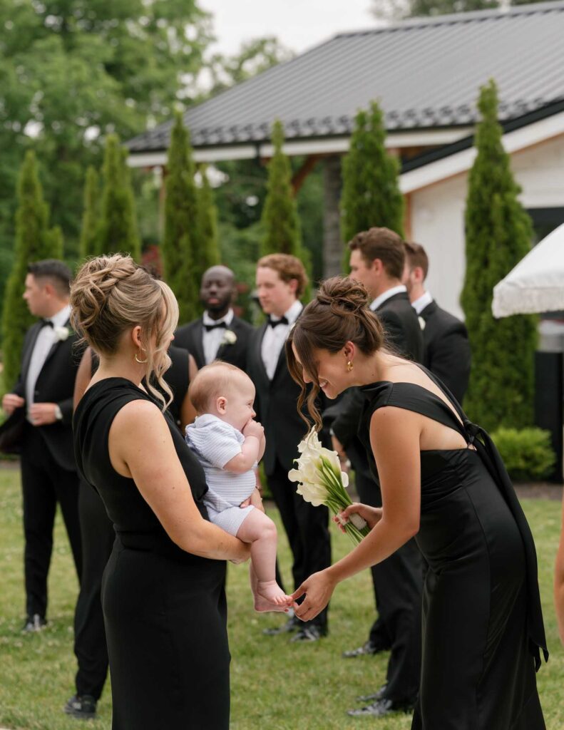A woman in a black dress holds a baby, while another in black kneels, smiling and holding flowers. Men in tuxedos converse in the background. The scene is joyful and elegant, set outdoors with trees and a building.