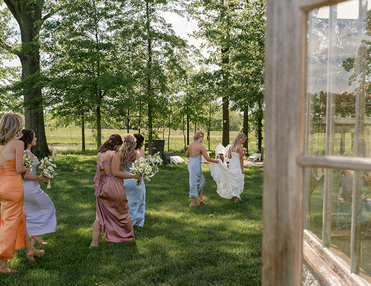 A bride in a white dress and bridesmaids in colorful gowns walk across a sunny, grassy field holding bouquets. Trees in the background add a serene touch.