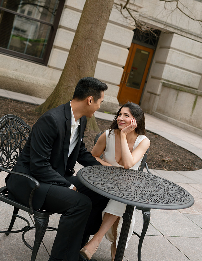 A couple sits at a metal table outside a building. The woman in a white dress smiles warmly at the man in a black suit, conveying a romantic mood.