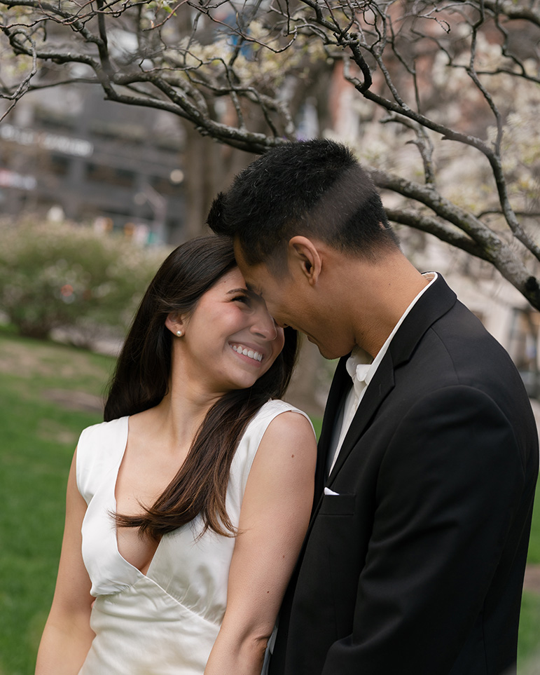 A couple in formal attire smiles warmly at each other under a blossoming tree, conveying joy and romance in a lush, green park setting.