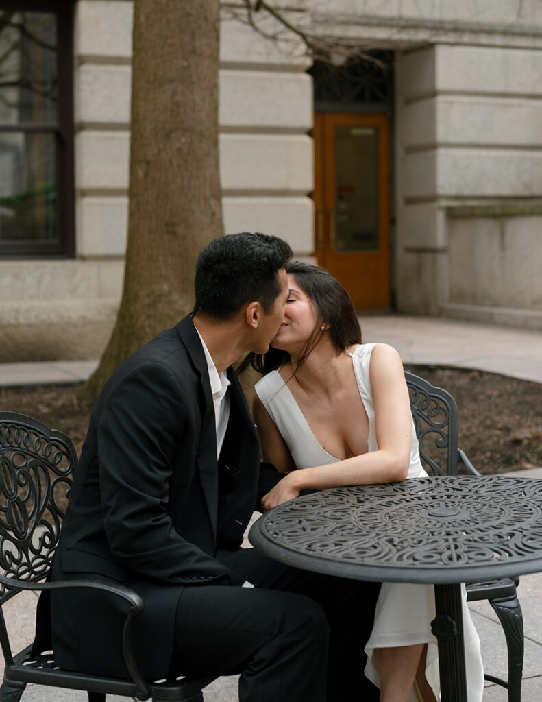 A couple kissing at a black wrought iron table in downtown Columbus, Ohio. The man wears a black suit, while the woman is in a white dress. The mood is romantic.