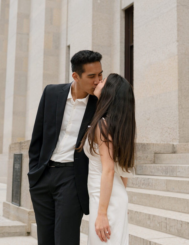 A Columbus couple in formal attire ascends stone steps beside large columns. The woman in a gown smiles back, holding hands with the man in a suit. Black and white photo.