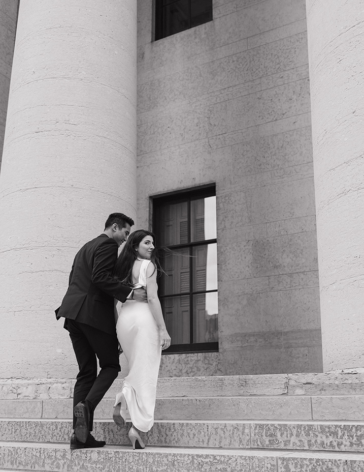 A couple elegantly ascends stone steps beside a grand pillar at the Ohio Statehouse. The man in a suit gently holds the woman in a flowing white dress, both smiling softly.