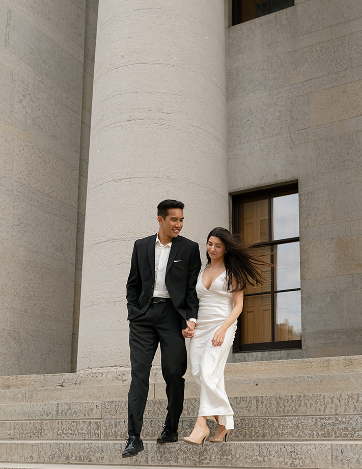 A couple walks down stone steps at the Columbus Statehouse holding hands in front of a large stone pillar. The man wears a black suit, and the woman is in a white dress, both smiling.