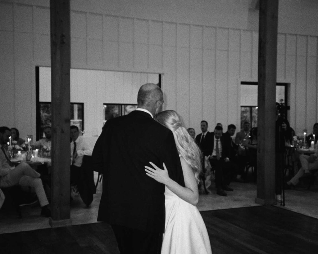 A bride and her dad share a tender dance on a dimly lit wooden floor, surrounded by seated guests, creating an intimate and heartfelt moment.