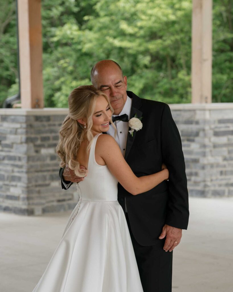 A bride in a white gown hugs an older man in a black suit, both smiling warmly. They stand under a wooden shelter with greenery in the background.