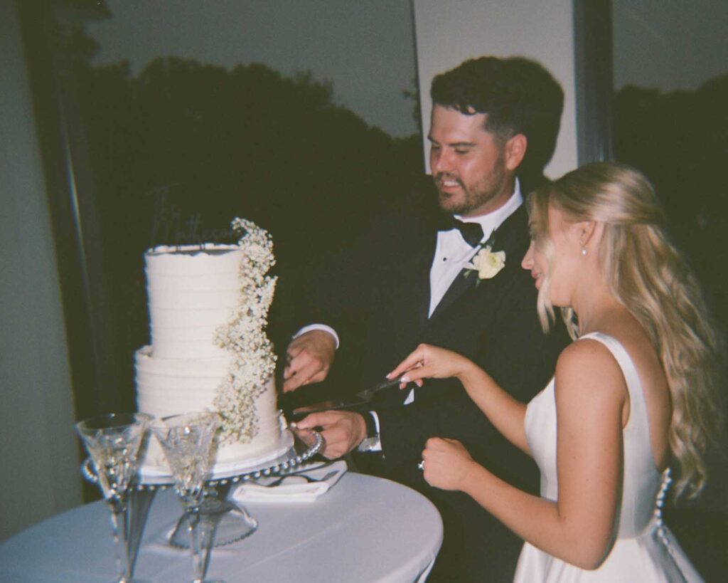 A bride and groom in formal attire cut a white, floral-adorned wedding cake together, smiling. Two champagne flutes sit on the table, adding a celebratory feel to the film style photo