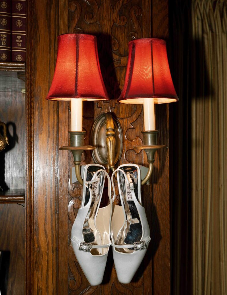 White high-heeled shoes with jeweled buckles hang on an ornate wooden wall sconce with red lampshades, giving a vintage and elegant feel.