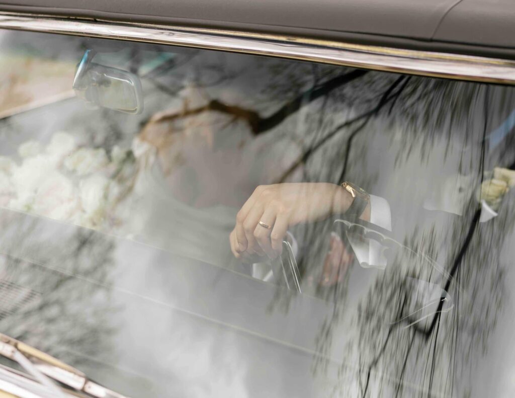 A hand with a wedding ring rests on a car steering wheel, visible through a windshield reflecting trees. The scene conveys calm and introspection.