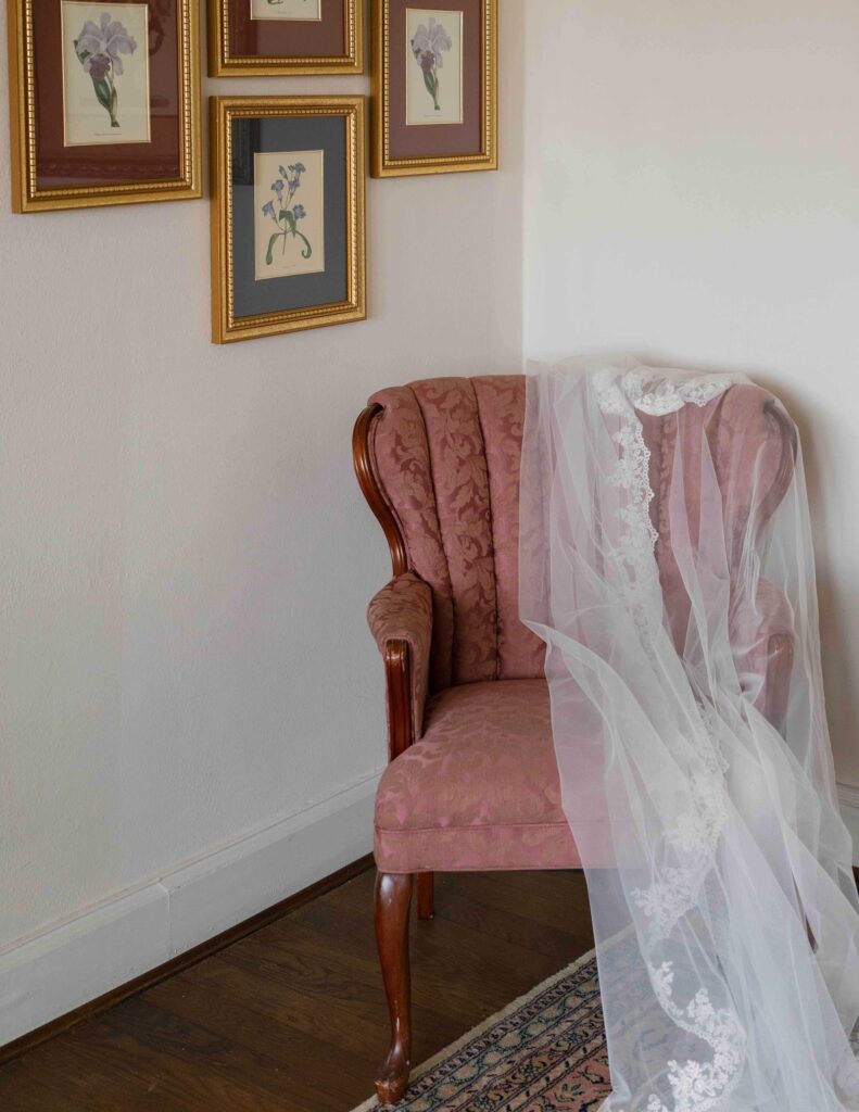 A delicate white lace veil drapes over a vintage pink armchair next to a wall adorned with framed floral prints, creating a tranquil, romantic ambiance.