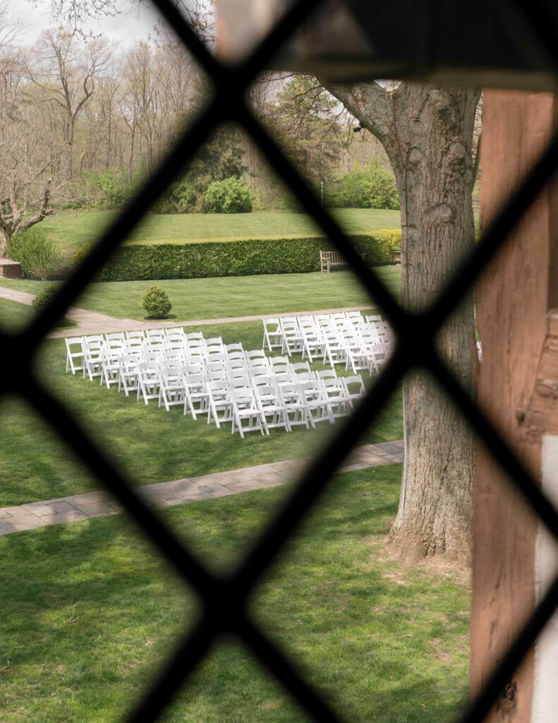 View through a lattice of an outdoor wedding setup, featuring rows of white chairs on green grass, framed by trees and a hedge in the background.