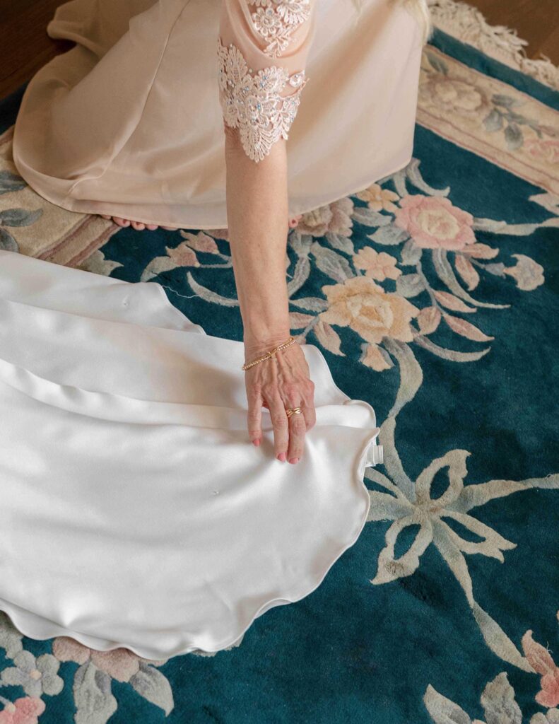 A woman in a light pink dress with lace sleeves adjusts a white satin fabric on a blue floral carpet. Her hand wears a gold bracelet, conveying elegance.