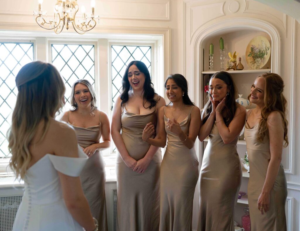 A bride in an off-shoulder gown stands smiling before five bridesmaids in champagne dresses. They express joy and excitement in a bright room.