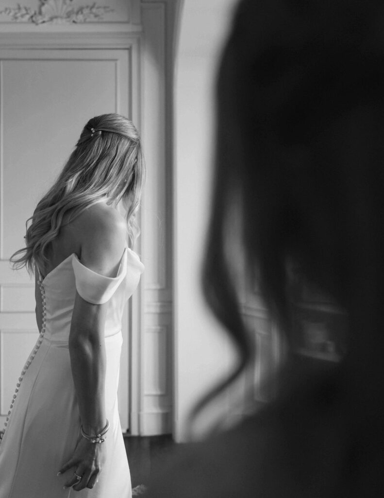 Black and white photo by Stevie J. Davis of a bride in a flowing gown facing away, her hair elegantly styled. A blurred figure appears in the foreground, adding depth.