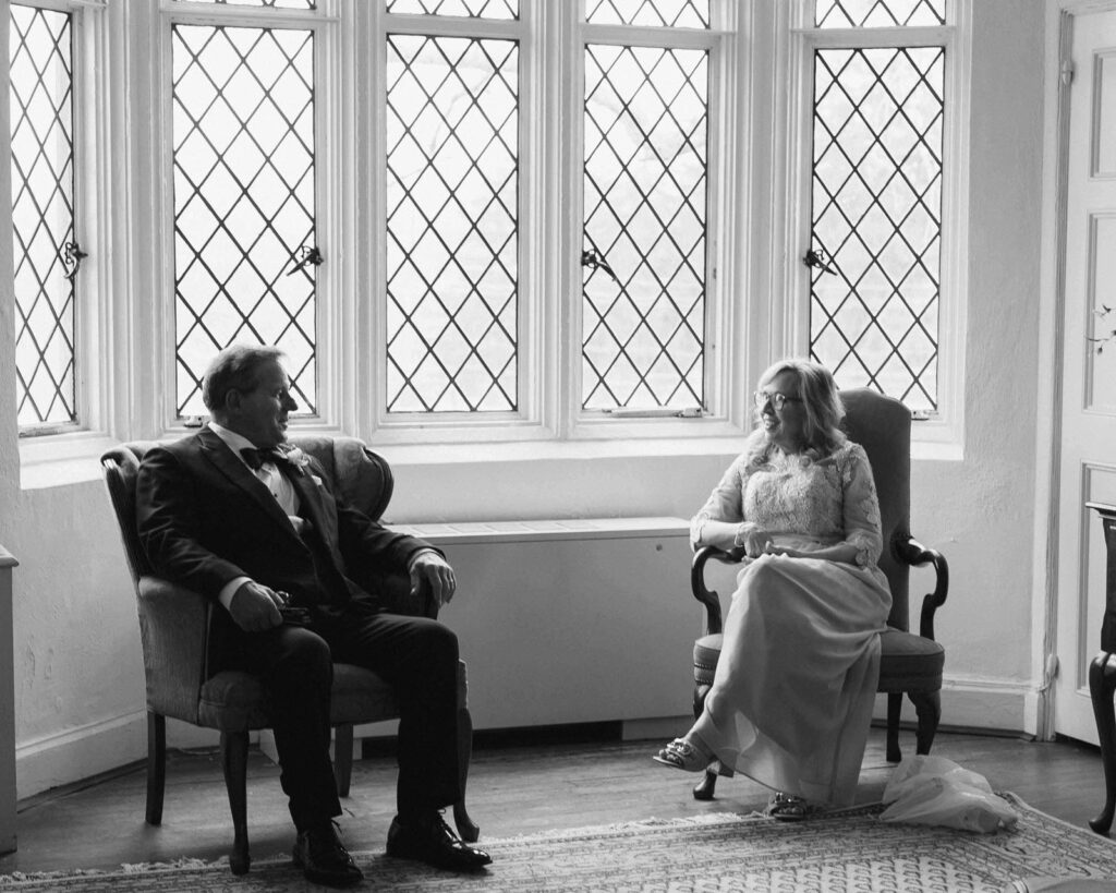 Black and white photo of a man in a suit and a woman in a dress seated on armchairs by large lattice windows, engaging in a relaxed conversation.