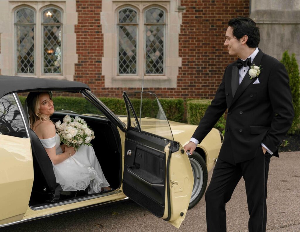 Bride seated in a vintage car holding white flowers, smiling at a tuxedo-clad groom holding the door. Romantic, elegant setting by a brick building.