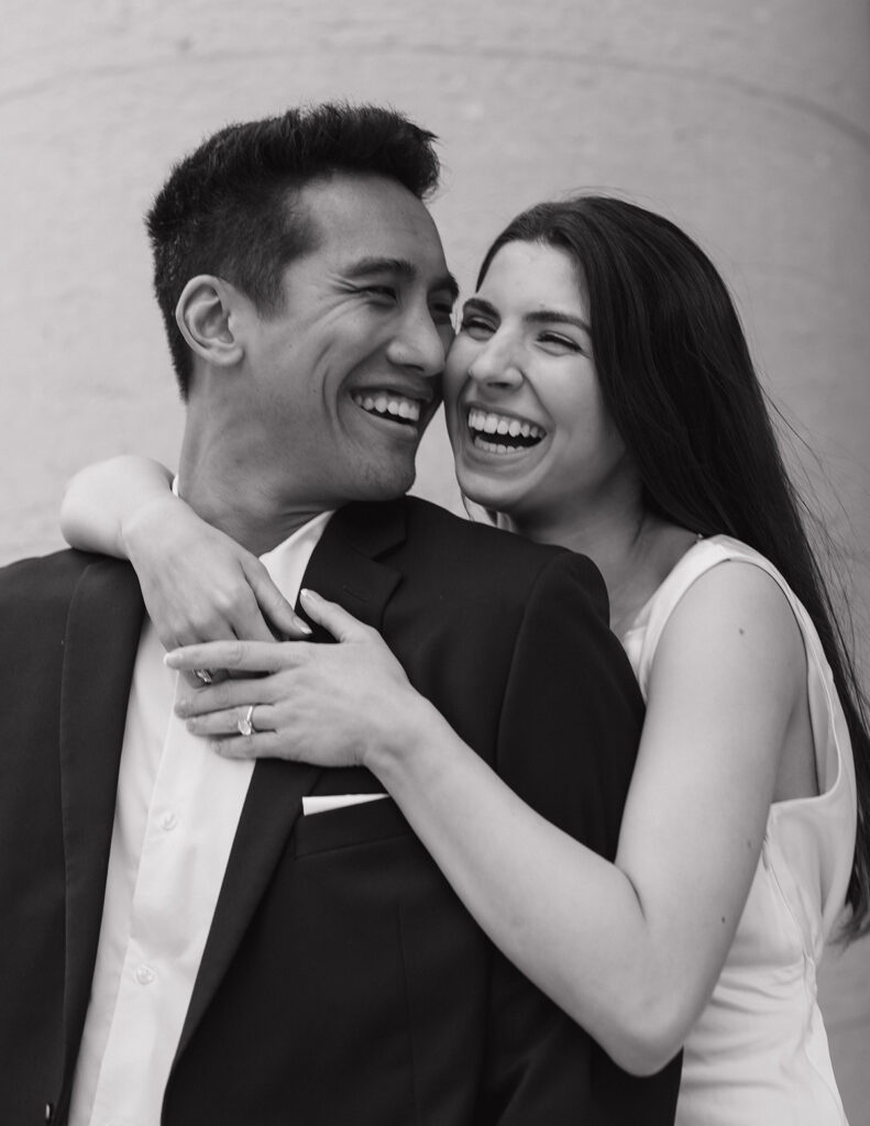 A joyful couple embraces, laughing warmly. The woman, in a sleeveless dress, leans on the man in a suit. The image conveys happiness and connection.