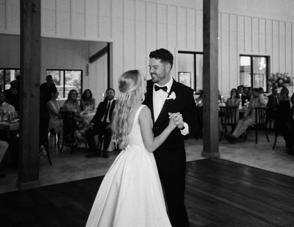 A couple shares a first dance at their wedding in a rustic venue. The groom, in a tuxedo, smiles warmly at the bride, who wears a flowing white gown. Guests watch happily.