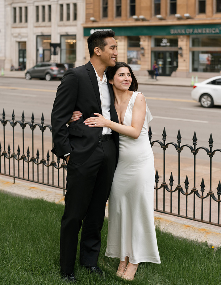 A couple stands close, smiling in elegant attire; the man in a black suit, the woman in a white dress. They're beside a wrought iron fence with downtown Columbus city buildings behind.