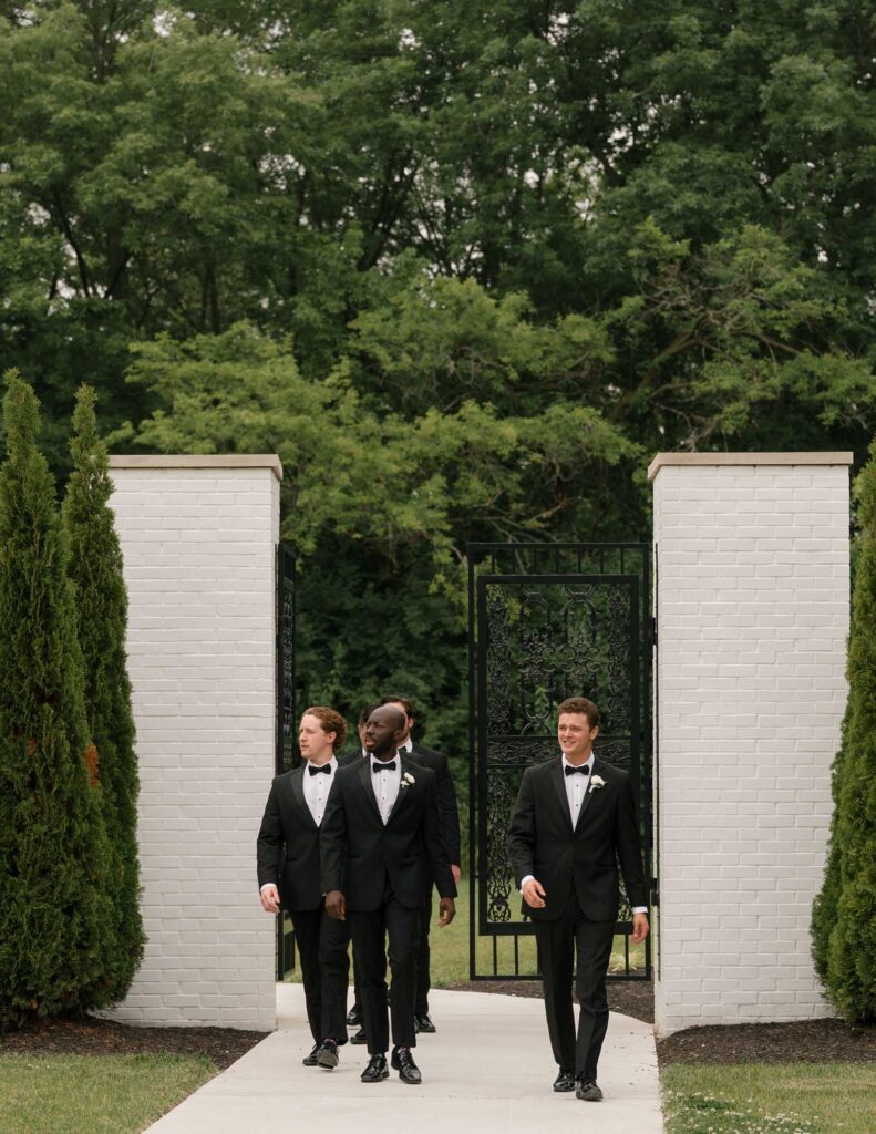 Four men in black tuxedos walk confidently through a garden gate. The scene is framed by lush greenery, creating an elegant and joyful atmosphere.