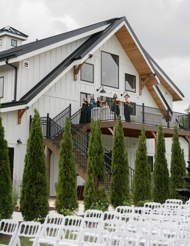 A group of people stand on a black-railed balcony of a white modern farmhouse, smiling. Tall trees line the foreground, and empty white chairs sit below.