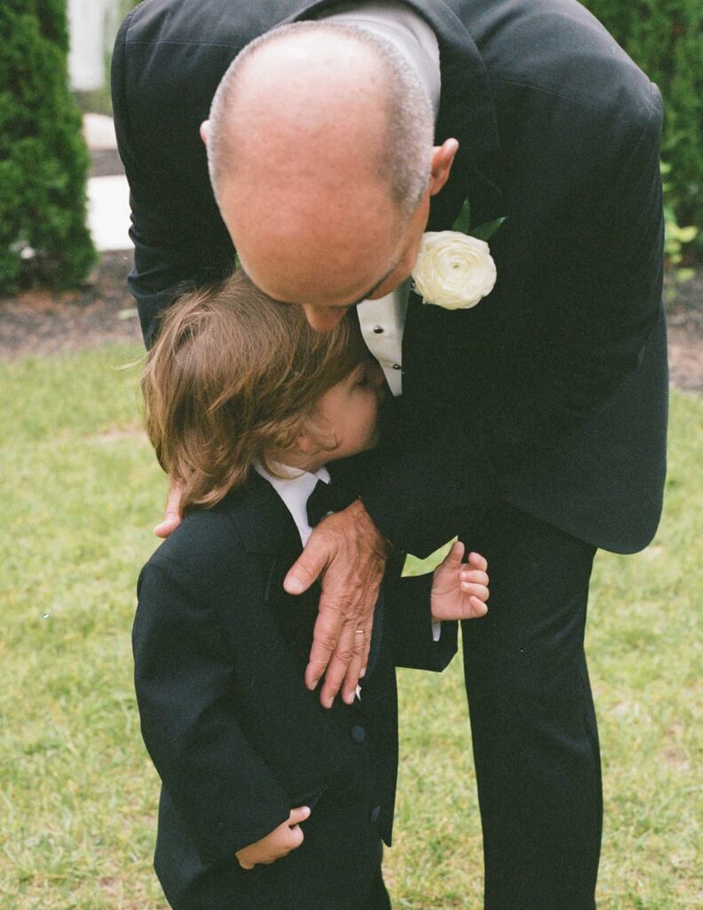 Film photo of a man in a black suit gently hugs a young child dressed in a matching suit, both appearing affectionate and tender, standing on green grass.