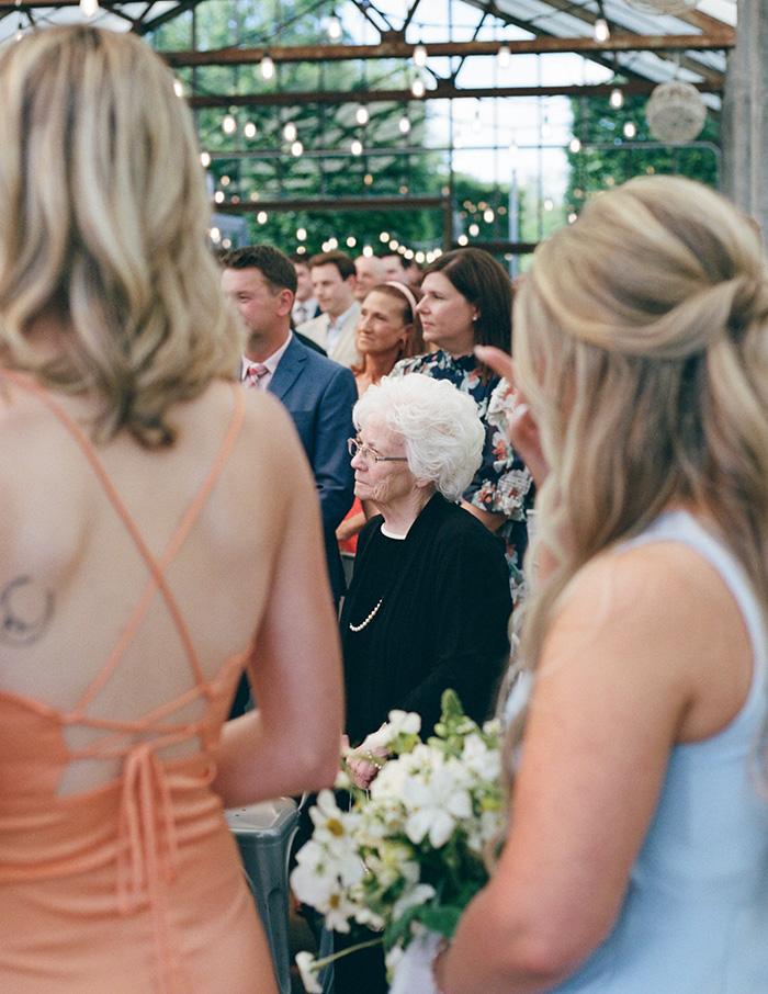 A diverse group of people, including an elderly woman in black, stand attentively in a decorated venue with string lights, evoking a formal event atmosphere.