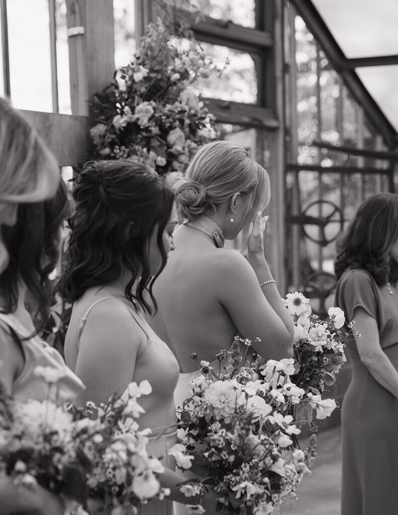 Bridesmaids holding bouquets in a glass-walled venue. One wipes a tear, conveying emotion. The mood is intimate and heartfelt. Black and white.