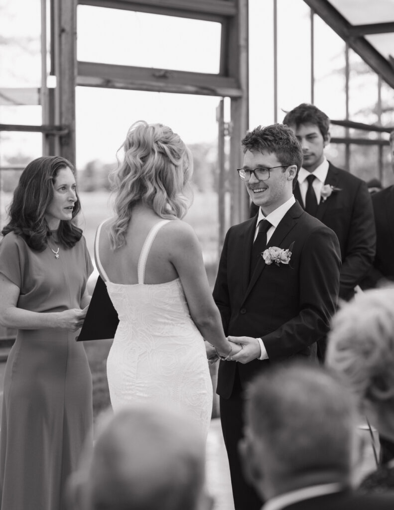 A bride in a white gown and a groom in a suit exchange vows, smiling, during an indoor ceremony. A woman officiates; attendees watch. Joyful atmosphere.
