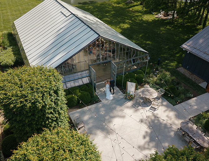 Aerial view of a glass greenhouse Oak Grove at Jergensen Farms with open doors, surrounded by lush greenery. A bride and groom are entering. The patio has string lights and tables. Peaceful ambiance.