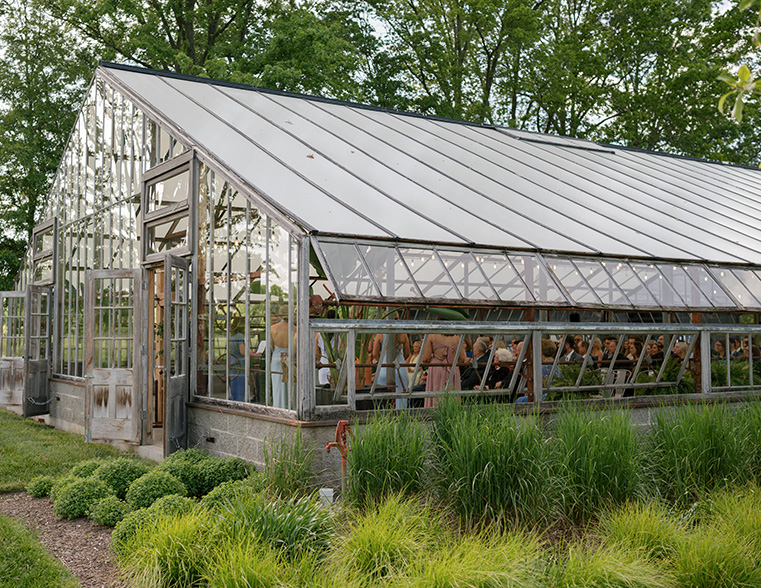 A glass greenhouse surrounded by lush greenery hosts a gathering of people inside. The scene is lively and illuminated, with trees visible in the background.