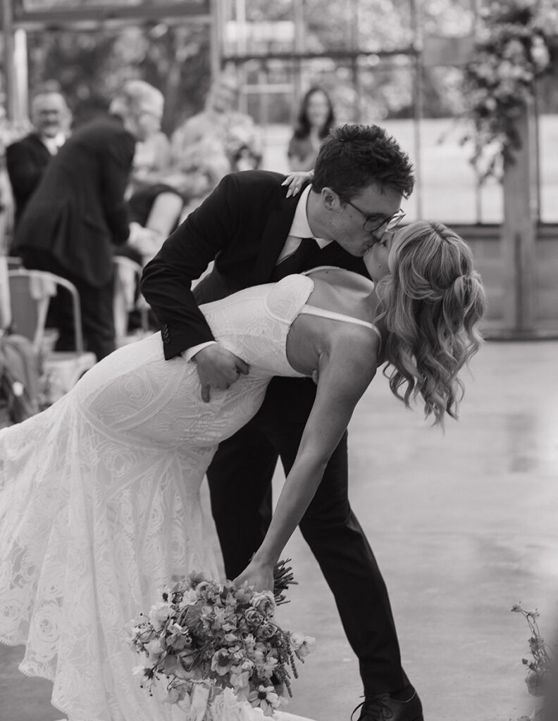 A bride and groom share a joyful kiss while the groom dips the bride, her white dress flowing elegantly. She holds a bouquet, guests smiling behind them.