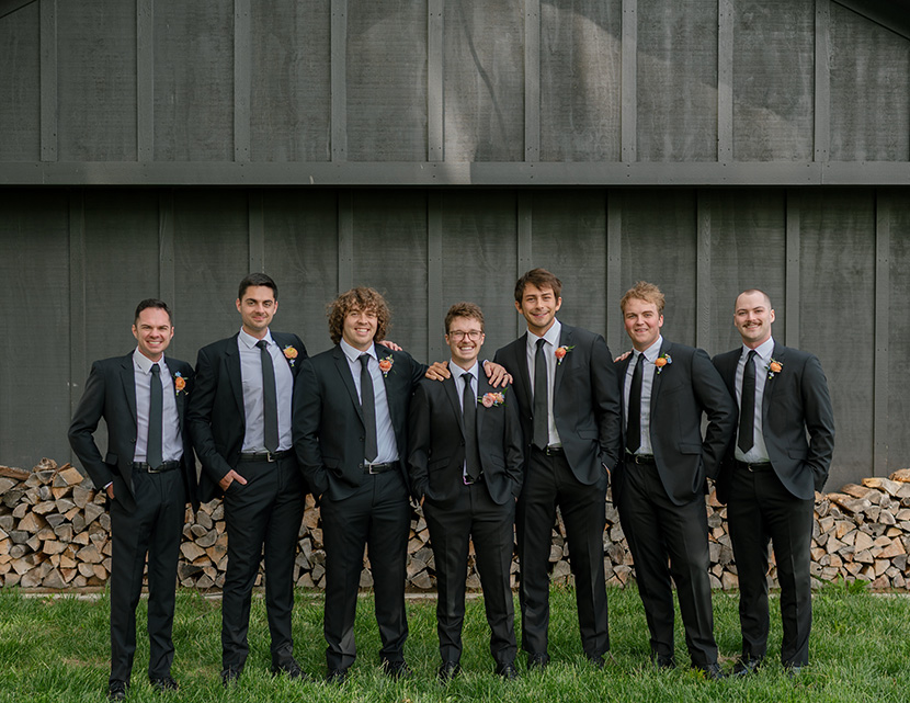 Seven men in matching dark suits and ties stand smiling in front of a wooden shed. They each have colorful boutonnieres, suggesting a wedding or formal event.