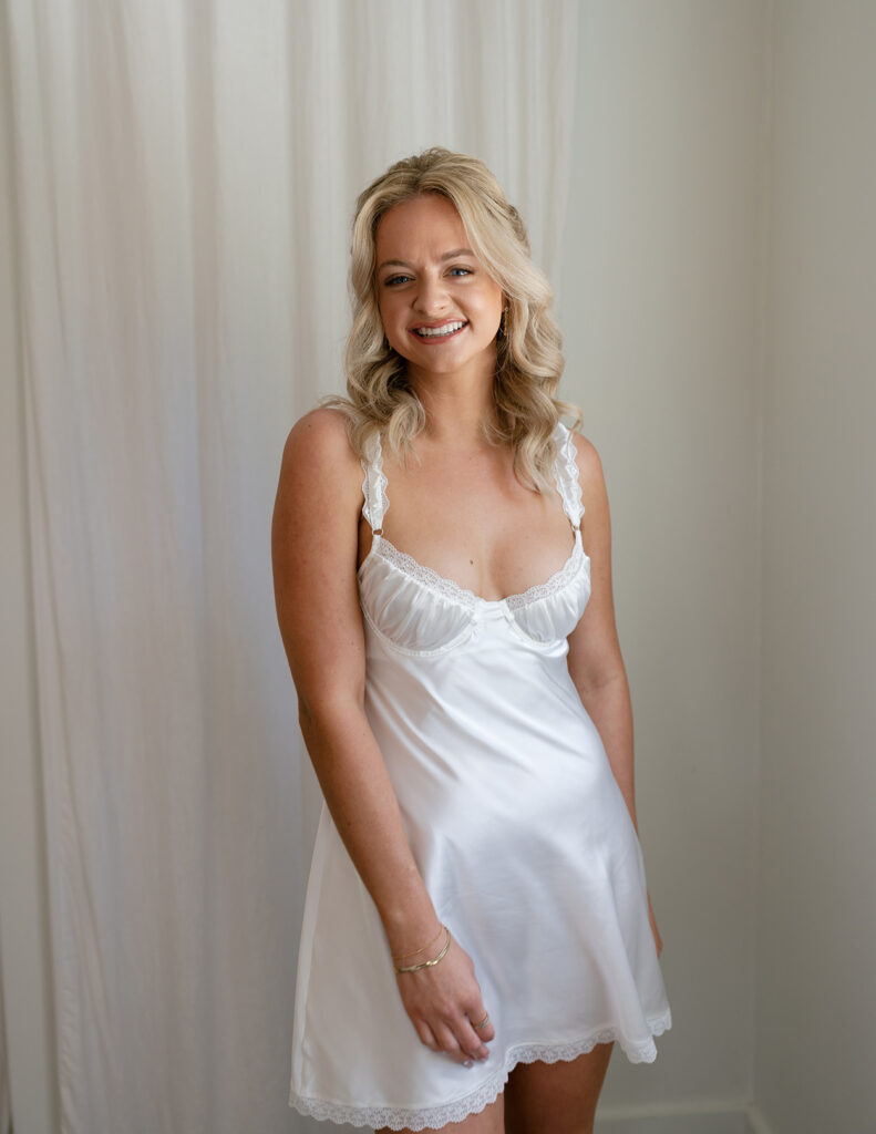A woman with blonde hair smiles warmly, wearing a white satin dress with lace straps, standing against a soft, beige background. The mood is cheerful and serene.