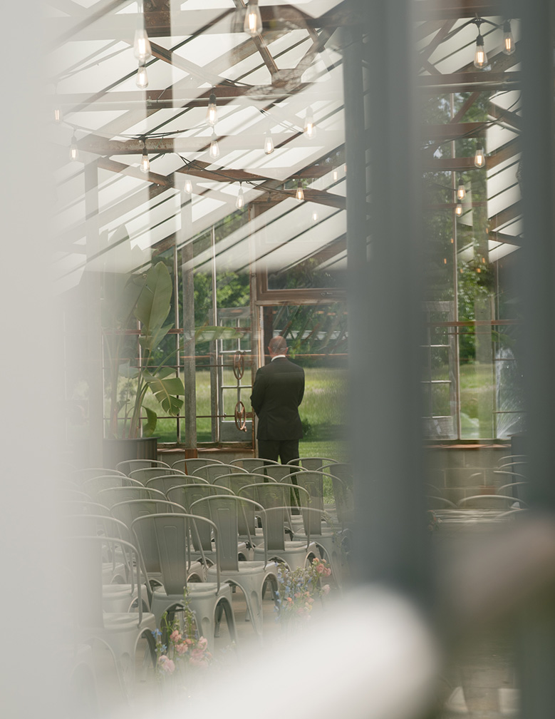 A man in a suit stands alone in Oak Grove Jorgesen Farms venue with rows of empty chairs and hanging lights. The mood is serene, with greenery visible outside.